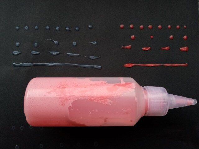 In the centre the transparent tube containing PVA glue mixed with red acrylic paint is shown. The upper left corners shows dry uncoloured glue, the upper right corner dry, red coloured glue. Compared to the colour of the mixture in the bottle, the dried